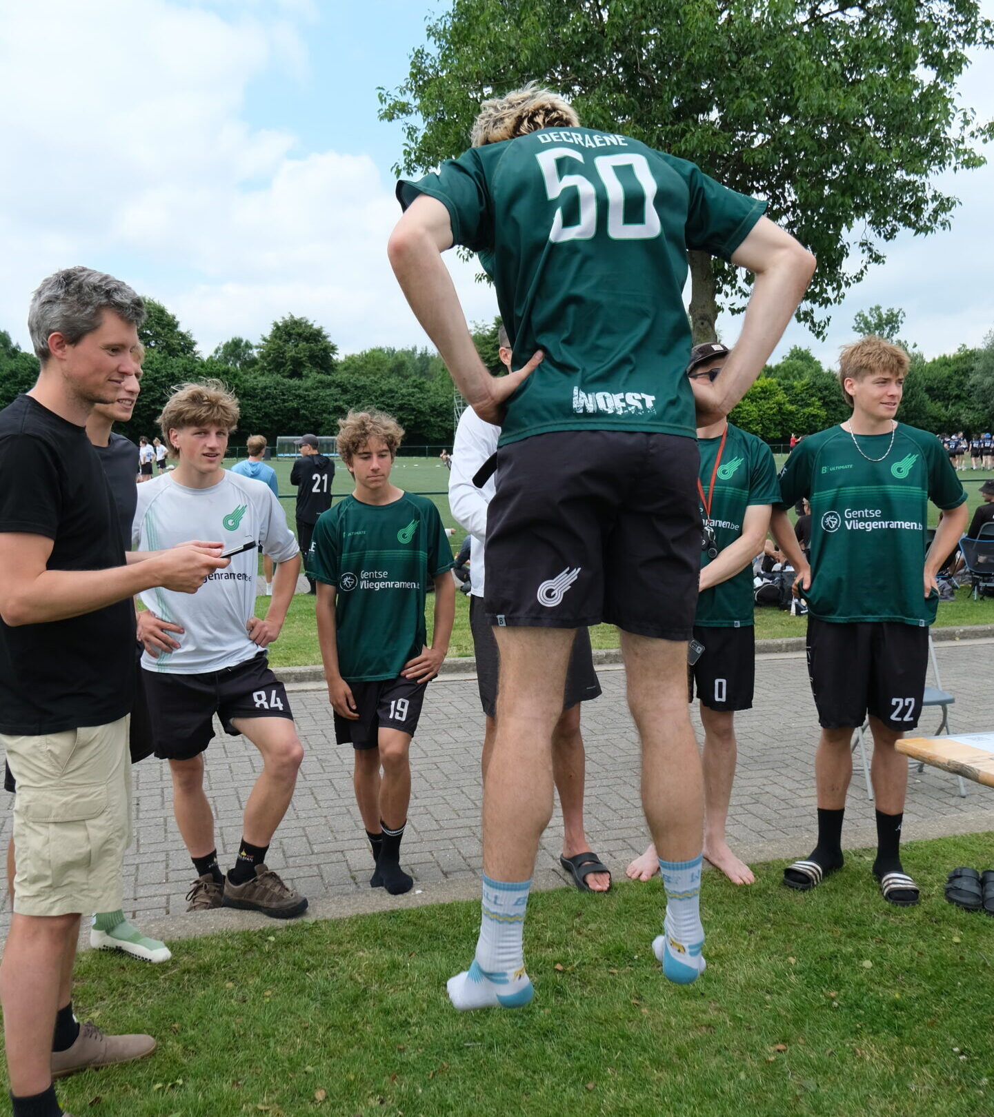 A group of young men in athletic gear are outdoors near a sports field. One man, wearing a green jersey labeled "Coppens 16," is captured mid-jump with his legs bent and feet off the ground, facing away from the camera. Another man to his left holds a tablet, seemingly measuring or observing the jump. Several other teammates in matching sports uniforms watch in the background. The setting is sunny with a partly cloudy sky, and the area is surrounded by greenery and sports equipment.